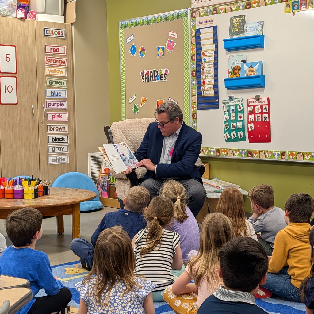 A man reads a book to a group of children in a classroom. They sit on the floor.