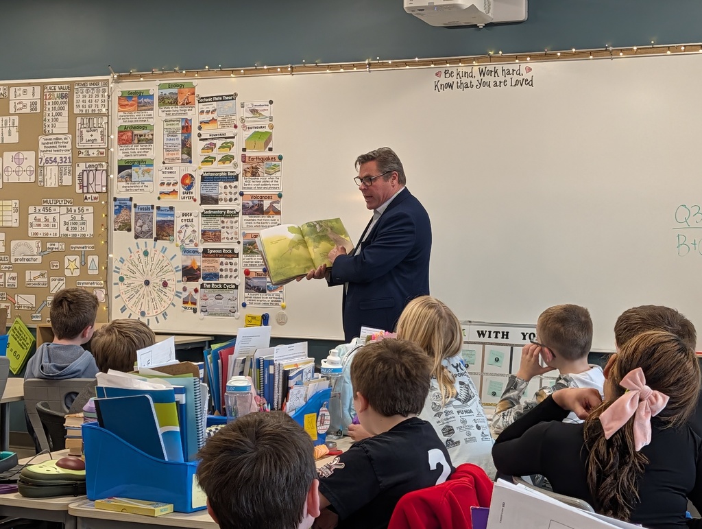 Man reading a book to a group of children seated around him in a classroom.