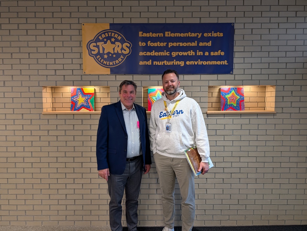 Senator Damoose and Principal Swegles stand next to each other in front of a blue banner and white brick wall.