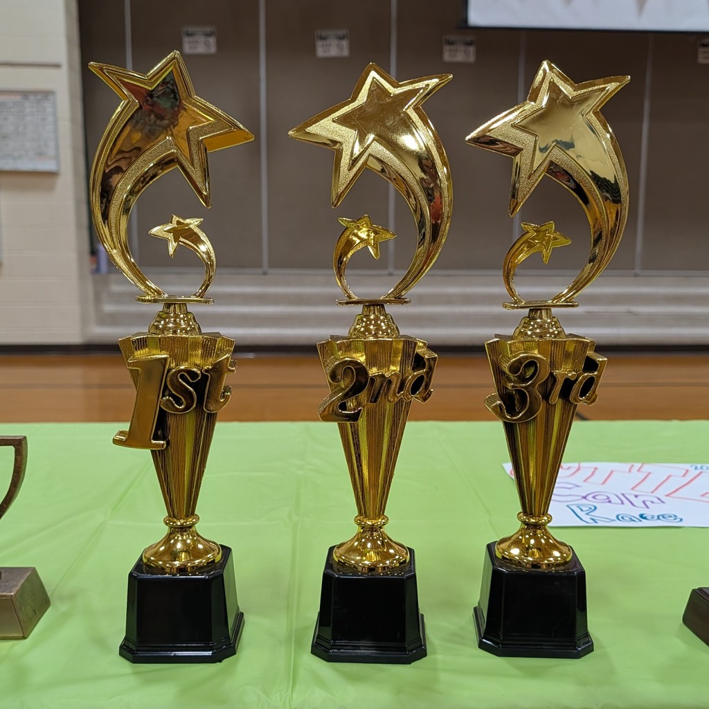 Three golden star trophies with black bases are lined up on a green table. The trophies are labeled 1st, 2nd, and 3rd.