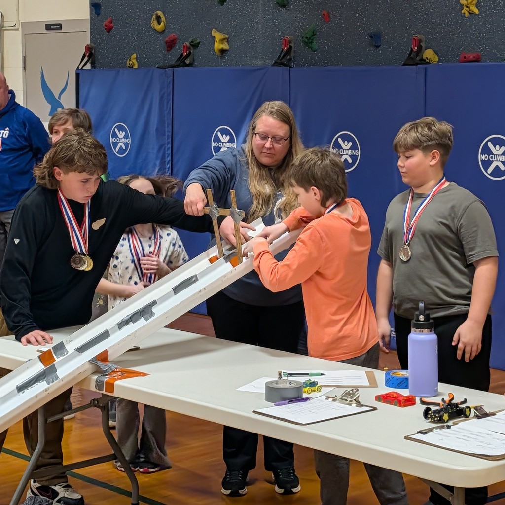 Students in a gym setting work on a project at a table, with a woman assisting.