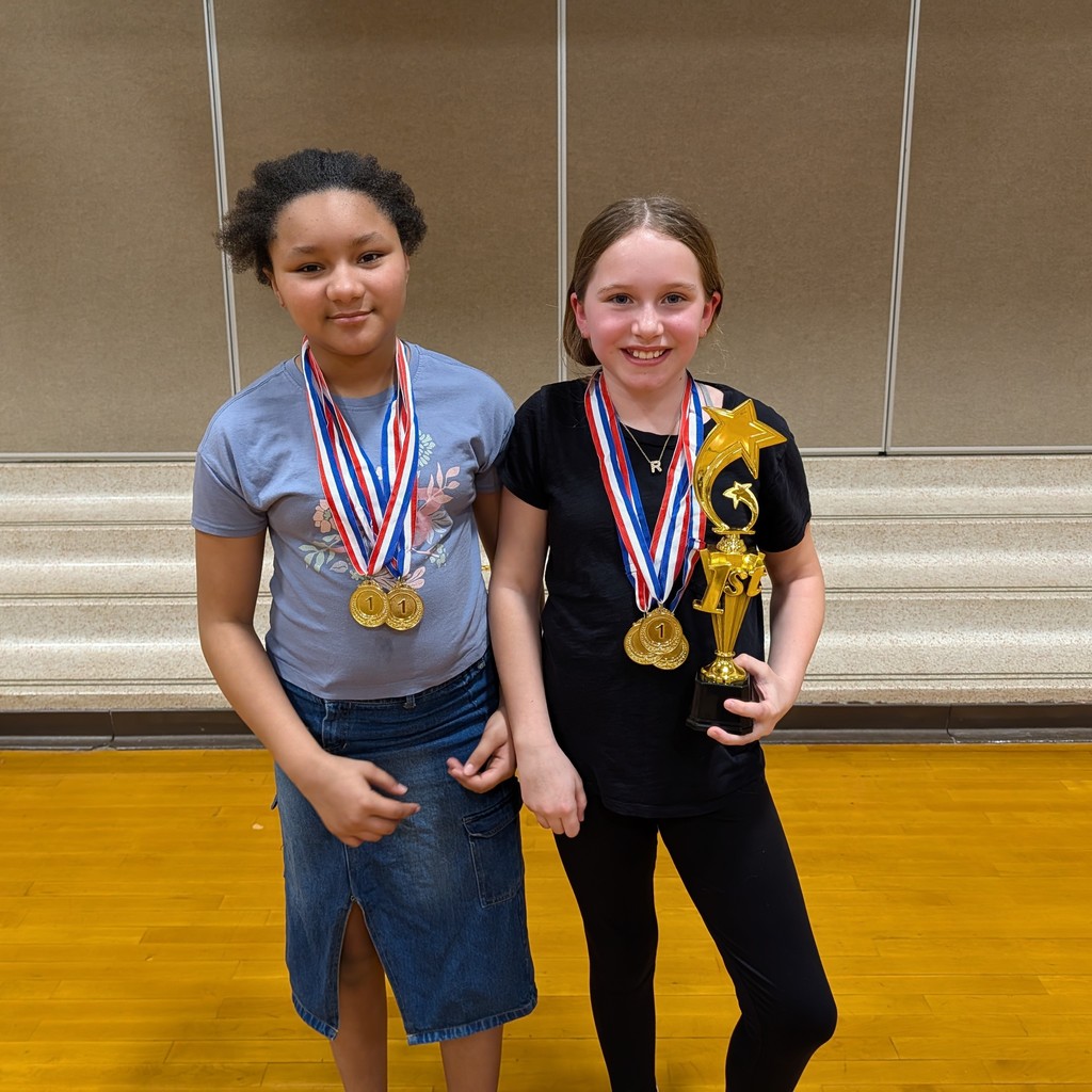 Two young girls stand next to each other holding medals and a trophy. One wears a gray shirt; the other, black.