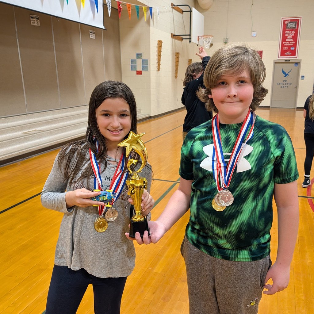 Two children with medals pose for a photo in a gym. One holds a trophy and medal, and the other has two medals around their neck.