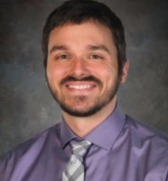 A man in a purple shirt and tie smiles with a mustache and beard.