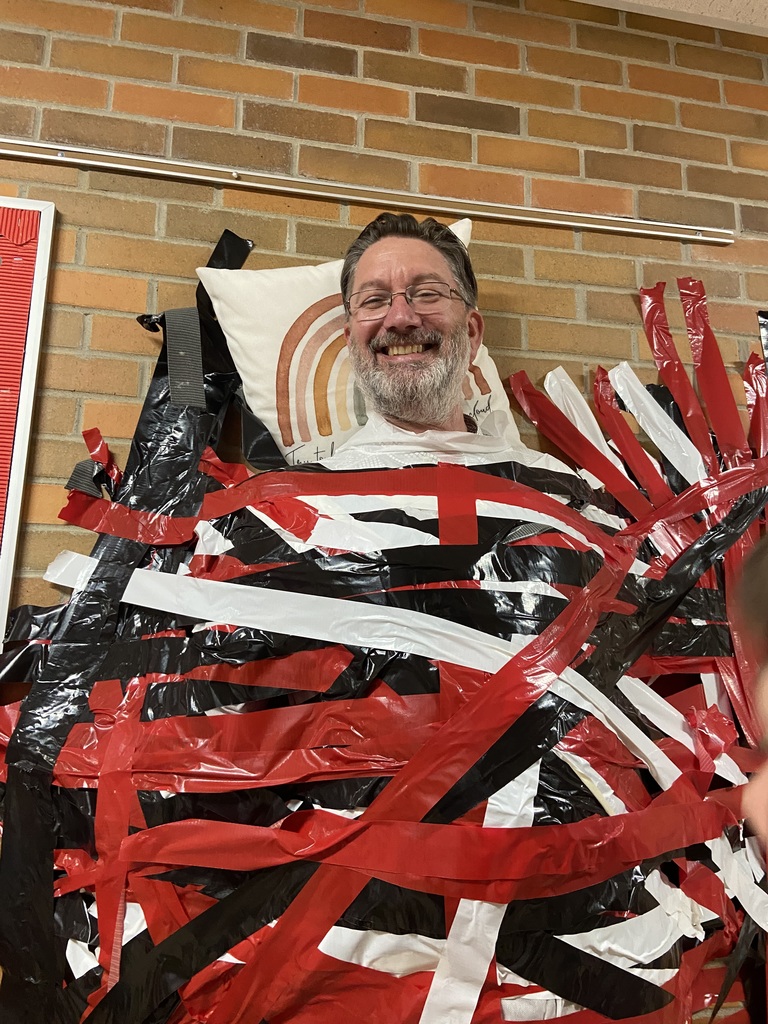 A Mr. Tisdale wearing glasses taped to the wall covered by red, white, and black plastic strips. Behind him is a brick wall.