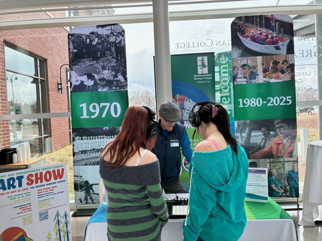 Two women in front of a table with banners. One banner shows the years 1970, and the other shows 1980-2025.