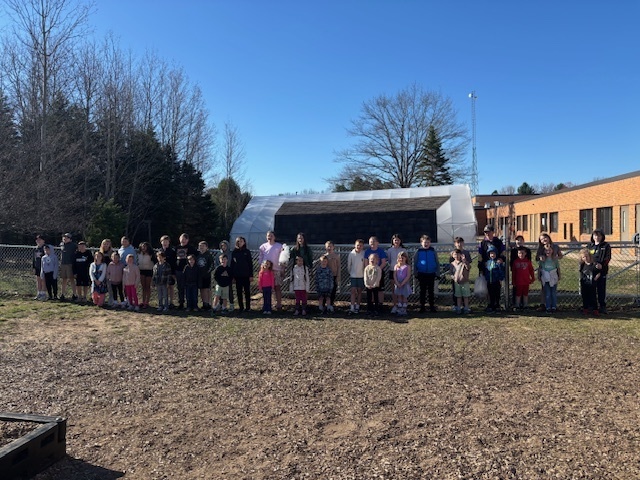 Many children pose in a field with a large memorial behind them. Trees and a building are in the background.