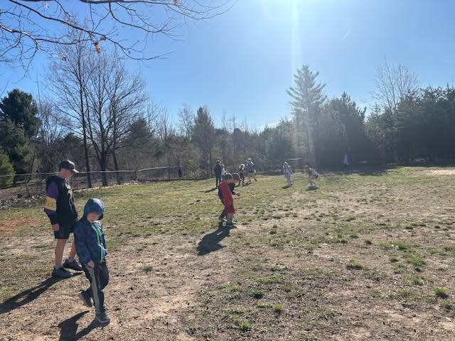 Several children walk on a dirt field with dry grass, surrounded by bare trees, under a clear sky.