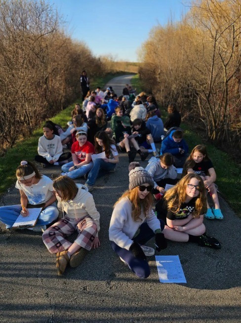 Many students sit on a path, likely for a field study. They hold papers and are surrounded by trees.