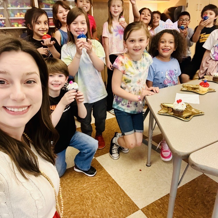 A group of children and adults smile in a room with cupcakes, tables, and a checkered floor.