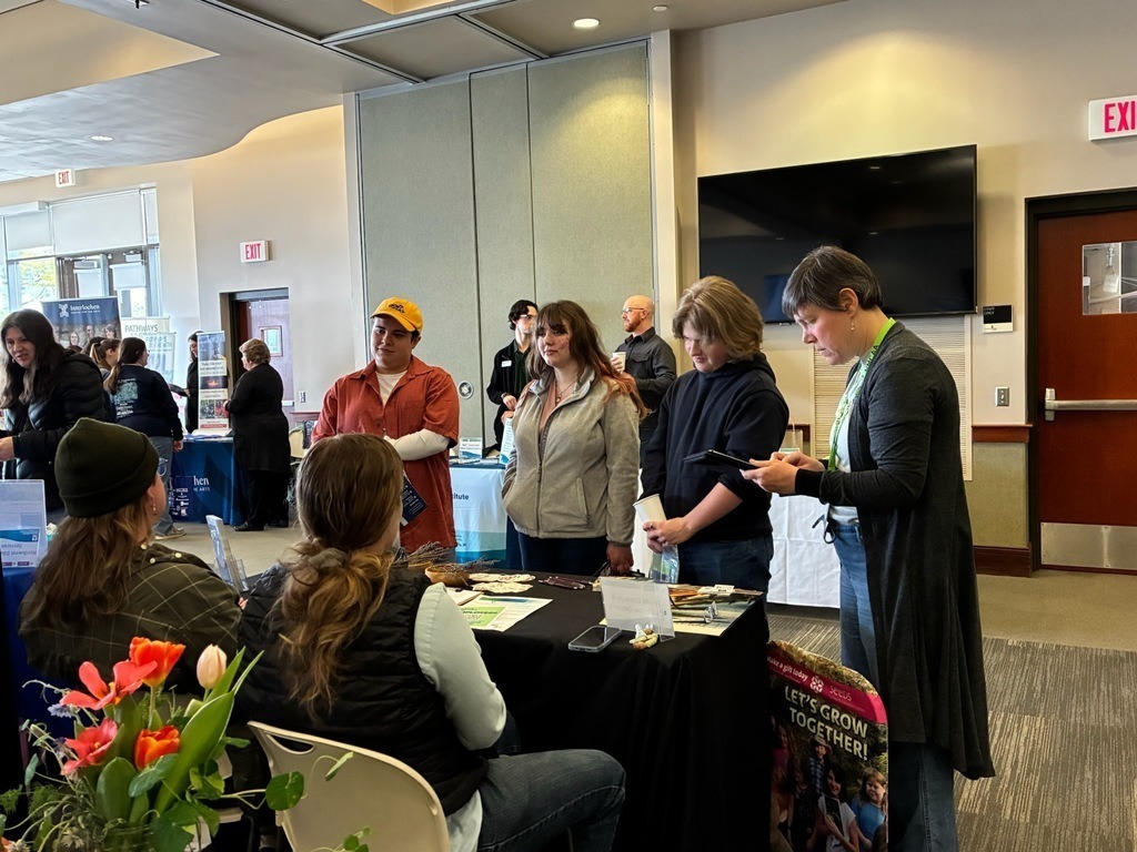 Several people are at a table with a woman, all looking at items. A TV is mounted on a wall.