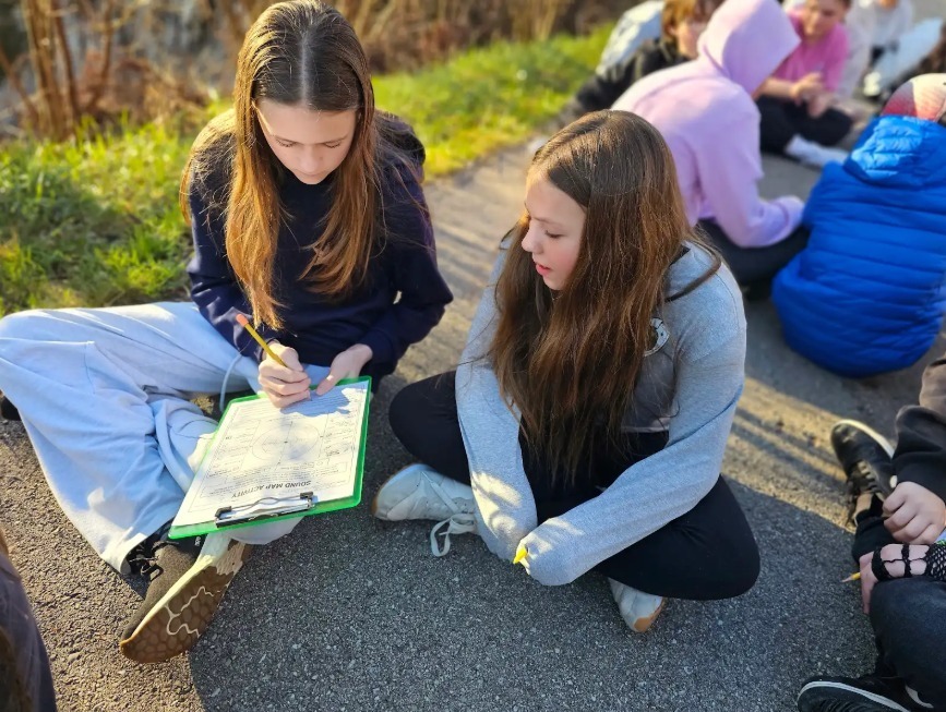 Two young women seated on the ground, one holding a clipboard and writing, the other holding a pencil.