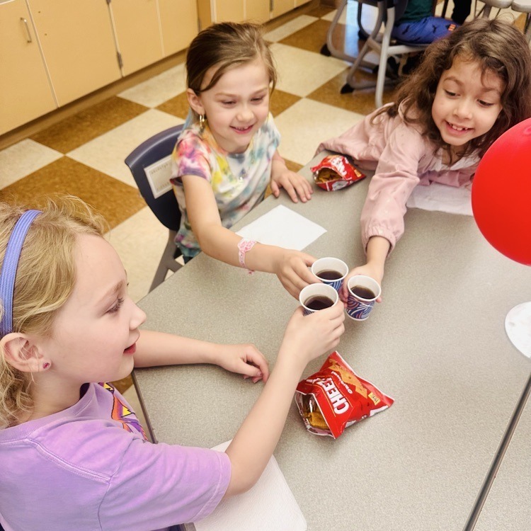 Three girls seated at a table hold cups, snacks, and a red balloon. The girl in a headband holds a cup.