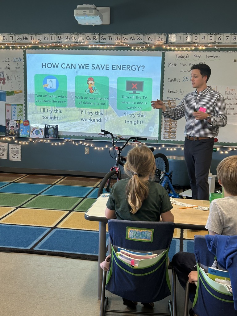 A teacher stands in front of a classroom screen showing an energy-saving presentation, with children seated around him.