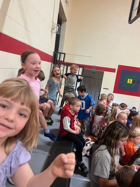 A group of children are in a school gym. Some are sitting on bleachers, others are standing.