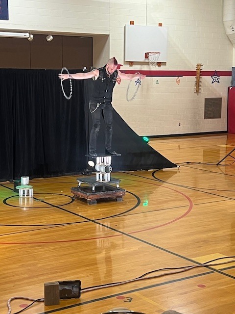 A person stands on a platform holding two hula hoops inside a gym with wooden floors.