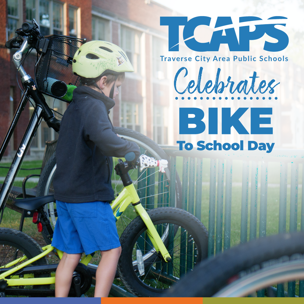 A boy with a helmet holds a bicycle, standing near a metal fence. A school banner reads "TCAPS Celebrates Bike to School Day."