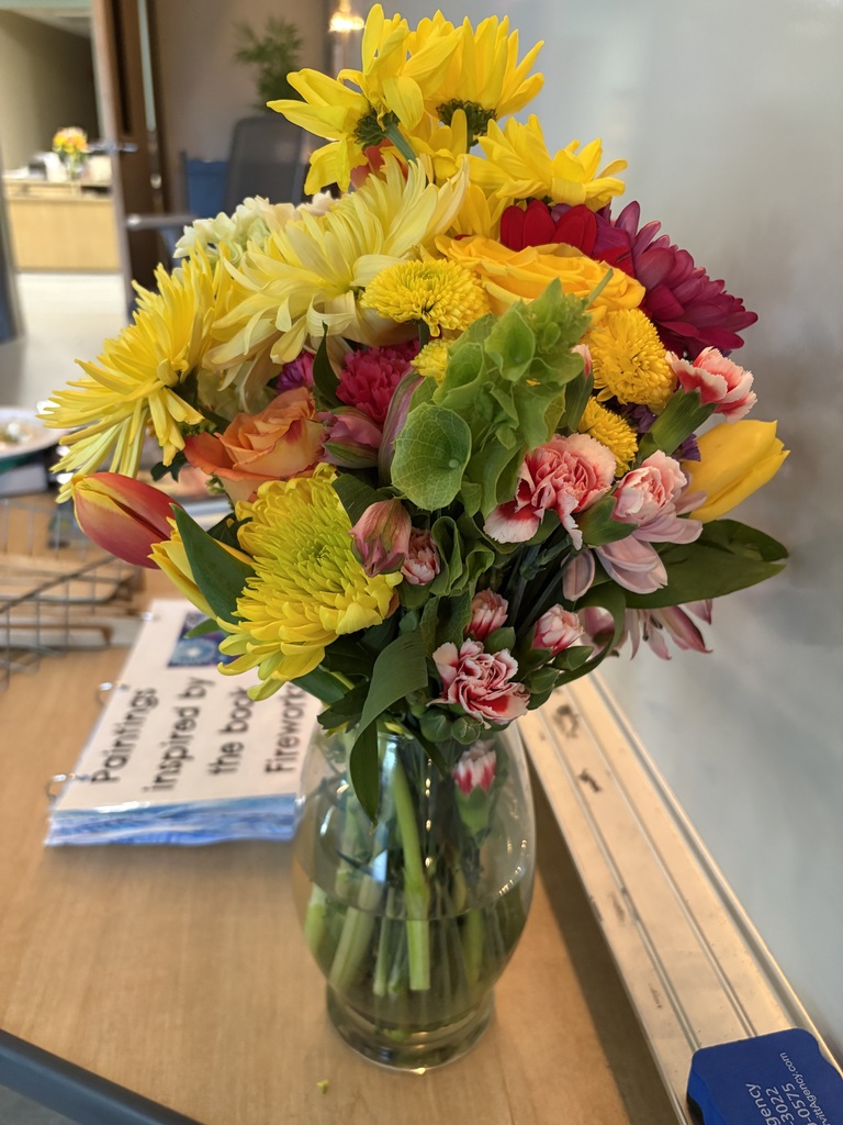 A vase with assorted colorful flowers, including yellow, red, and pink, on a wooden table. 