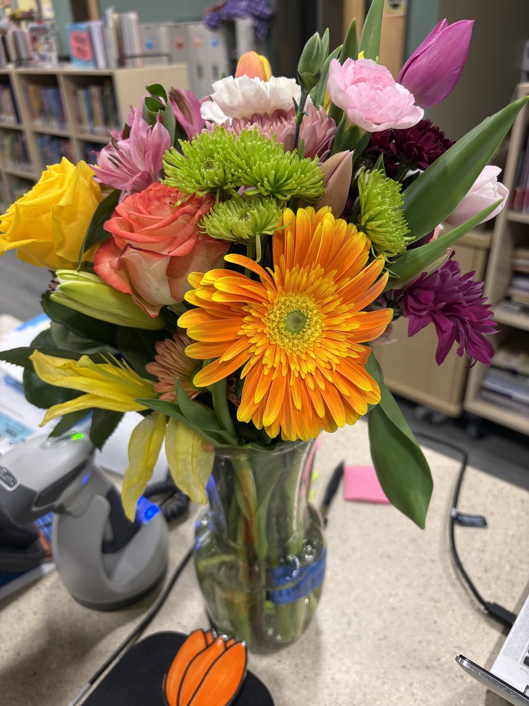 Colorful flowers in a glass vase on a table. Behind the vase is a book shelf.