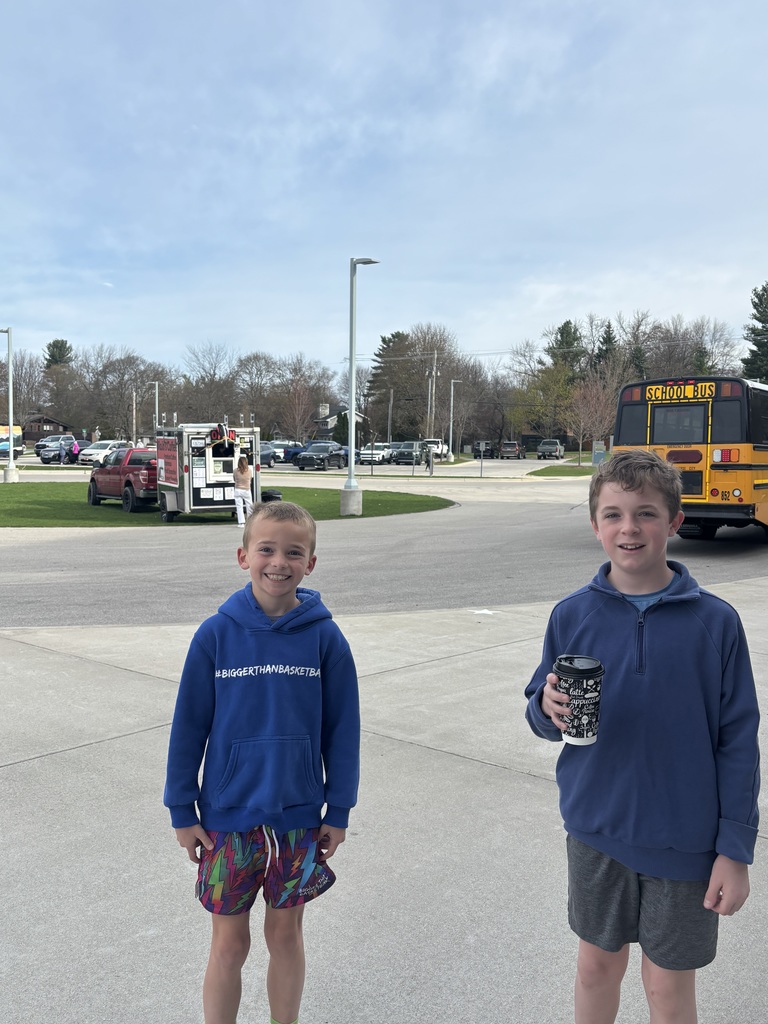 Two young boys stand in a parking lot. One holds a cup, and a school bus is in the background.