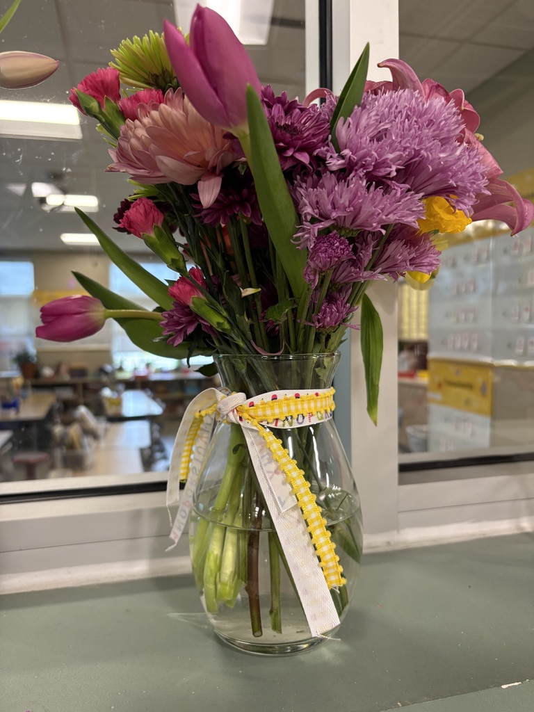 A clear glass vase with a ribbon holds a bouquet of purple and pink flowers in front of a window.
