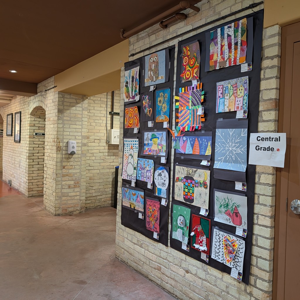 Hallway with brick walls. A bulletin board displays various artworks. One sign reads "Central Grade."