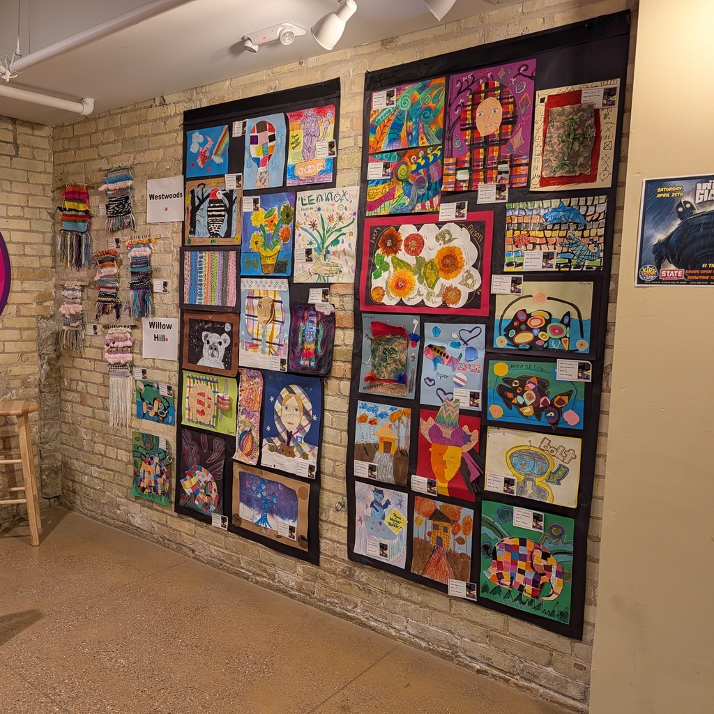 Art pieces in frames adorn a brick wall. Brightly colored paintings are displayed. A stool is on the left.