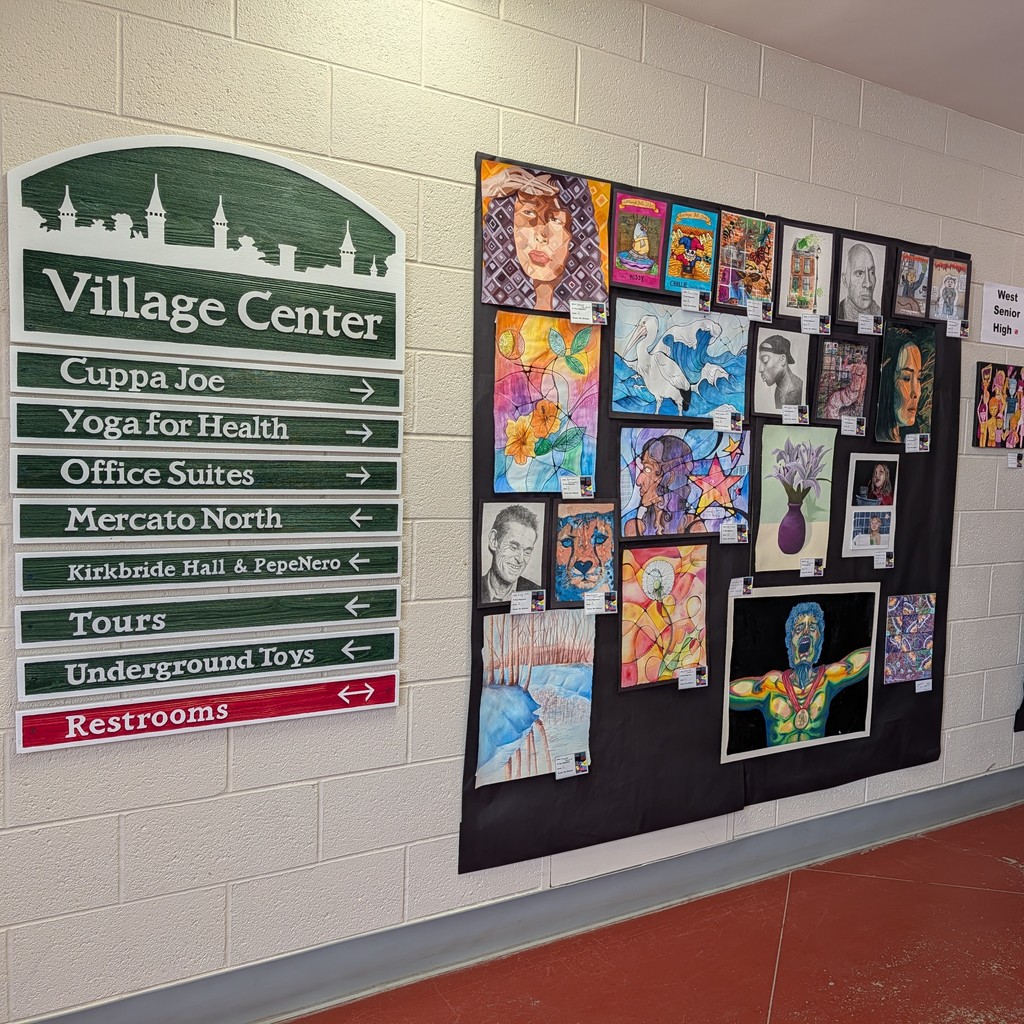 A white wall with a sign pointing to various locations and an art display with multiple colorful paintings.