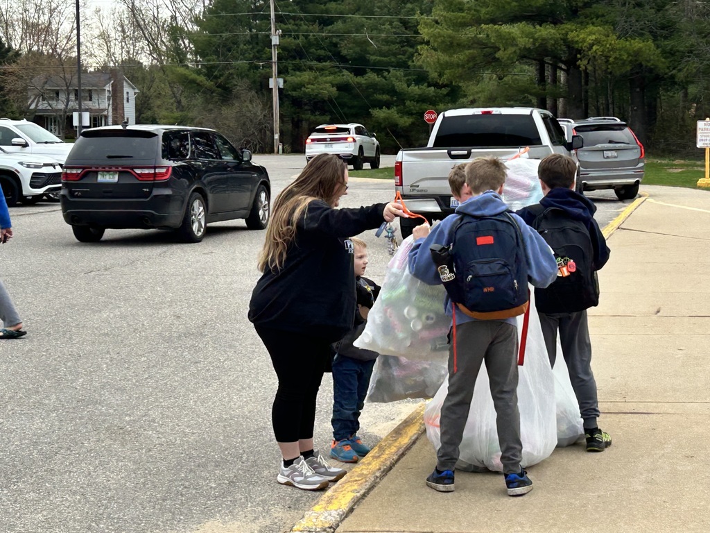 A woman assists a group of children, who carry bags, on a sidewalk. Cars are passing by.
