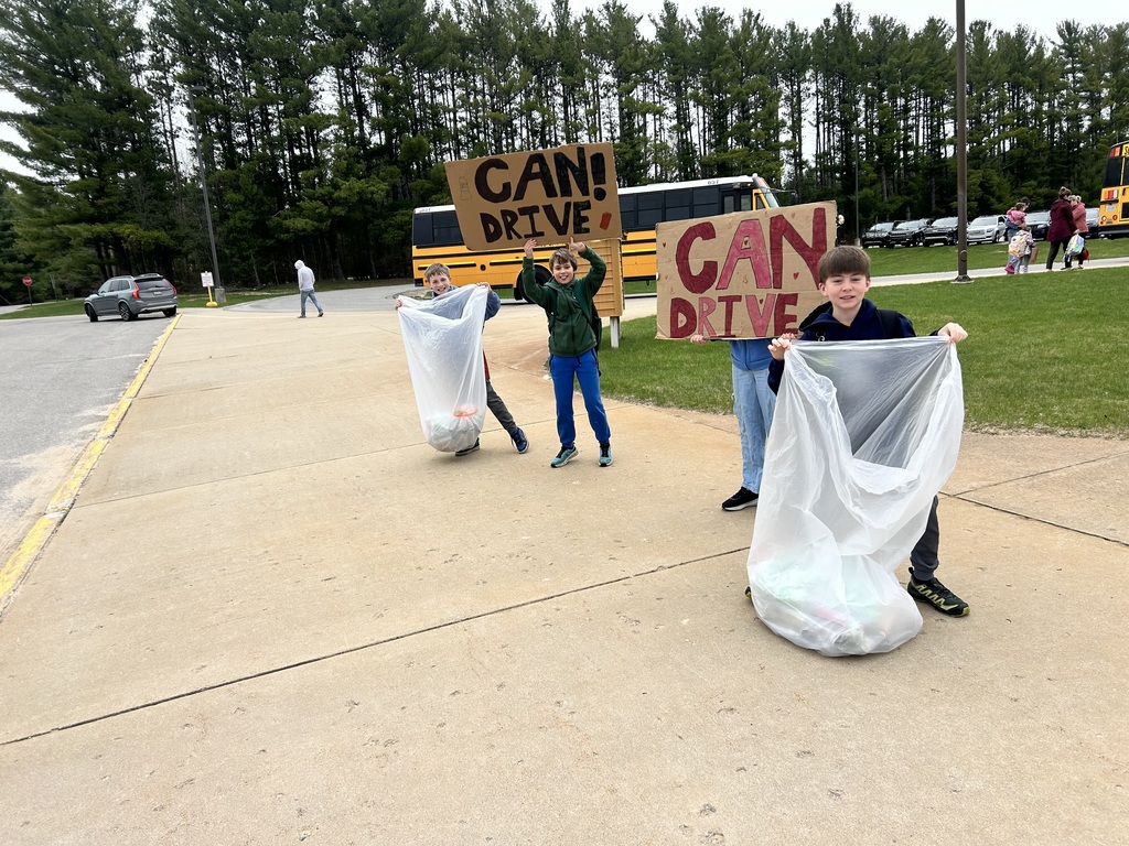 Four people hold signs reading "CAN! DRIVE" while holding bags. They stand on a sidewalk with a grassy area behind them and trees and a bus in the background.