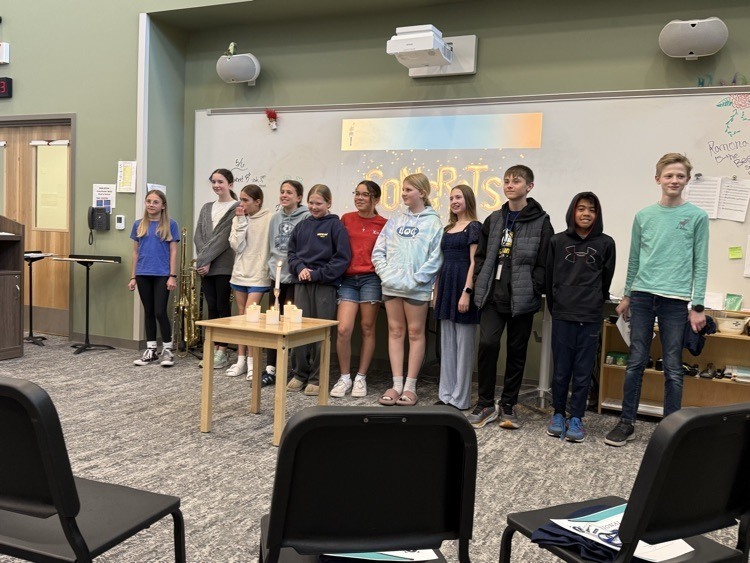 A group of students stands in a room with a wooden table, chairs, and a whiteboard in the background.