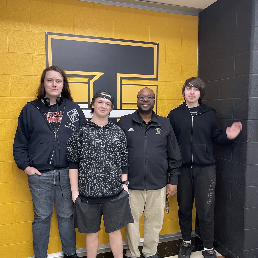 Four people in dark clothing stand in front of a yellow wall with a black and yellow logo.