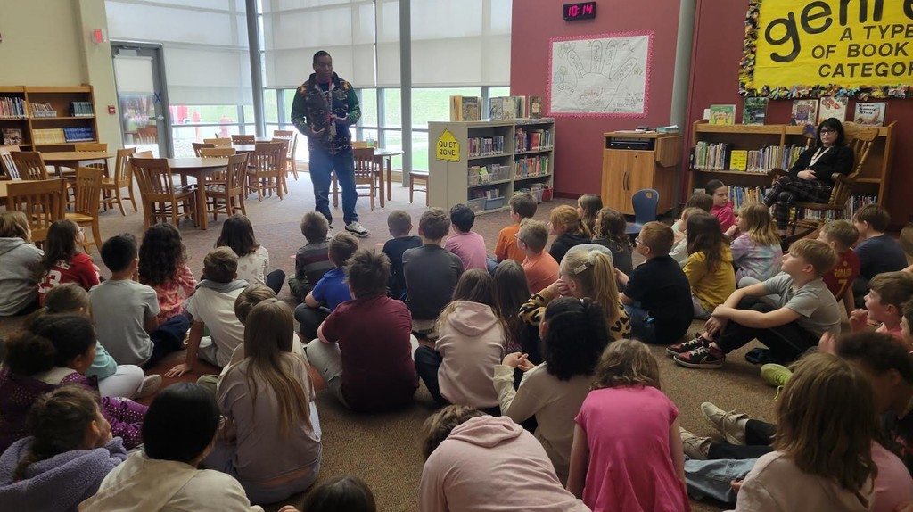 A guest speaker in a patterned jacket speaks to a group of children seated on a library floor.