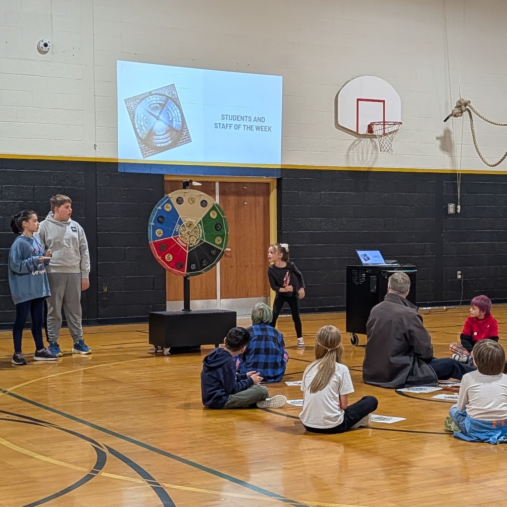 Several students sit in a gymnasium, facing a large spinning wheel. A projector screen displays behind the wheel.