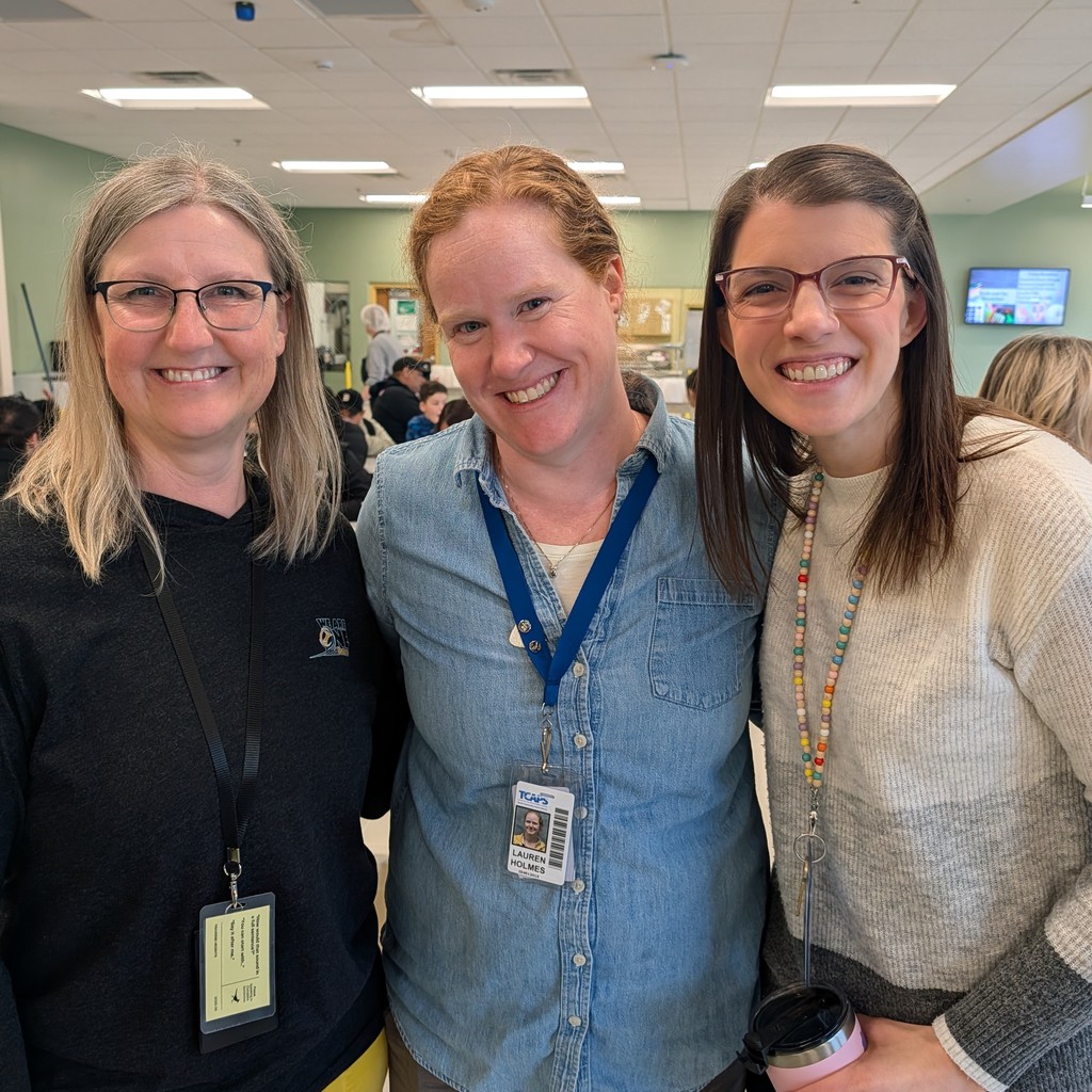 Three women in a room with white walls and ceiling lights, smiling. Two wear glasses. The one in the middle wears a blue shirt and ID.