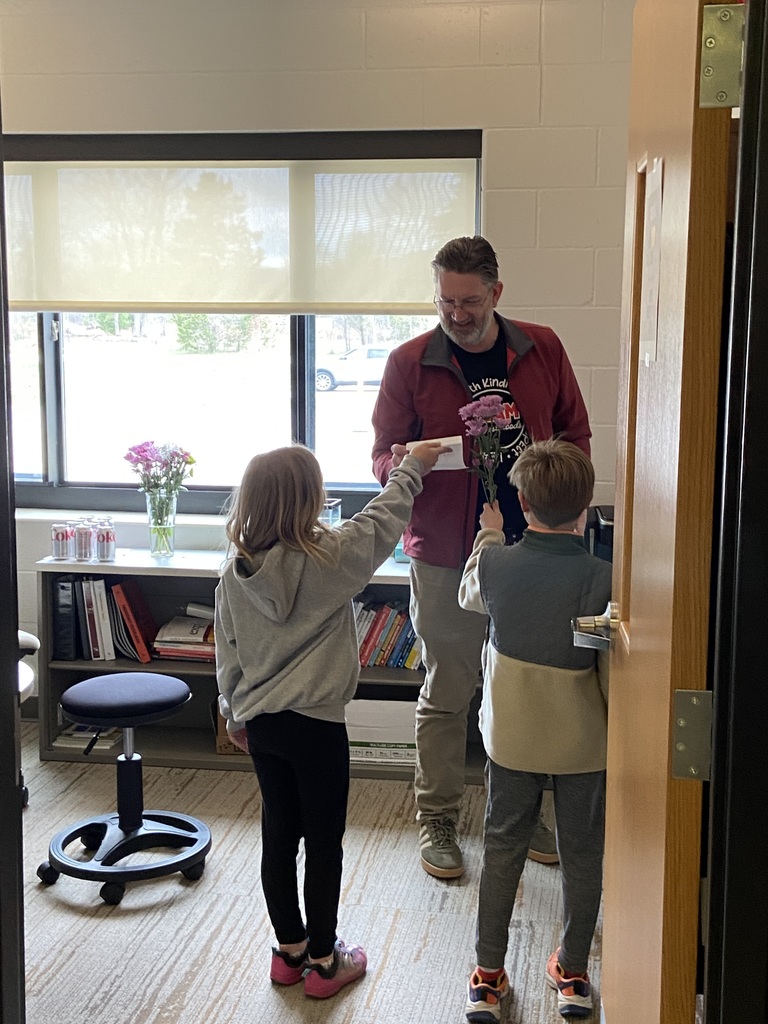 A man and two children inside a room with a bookshelf, a flower vase, and a window with blinds.