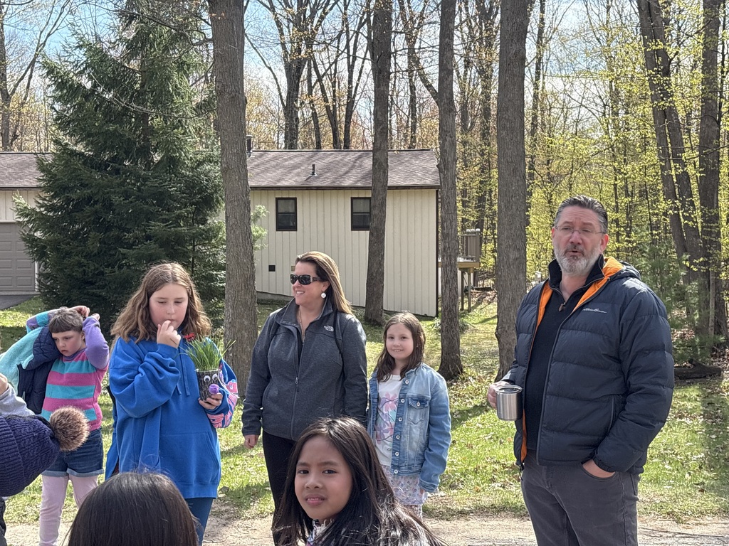A group of children and adults stand outside near a house. Trees are visible. A man stands with his hand in his pocket.