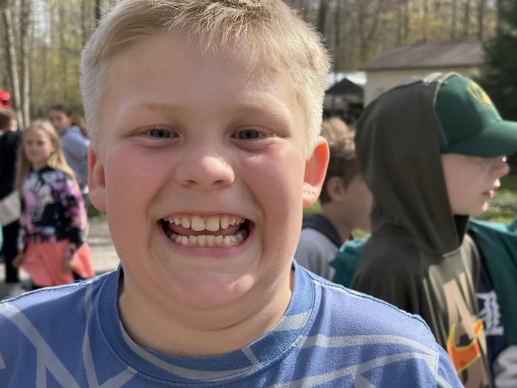 A boy with blonde hair smiles widely in a blue shirt, with other children and trees in the background.