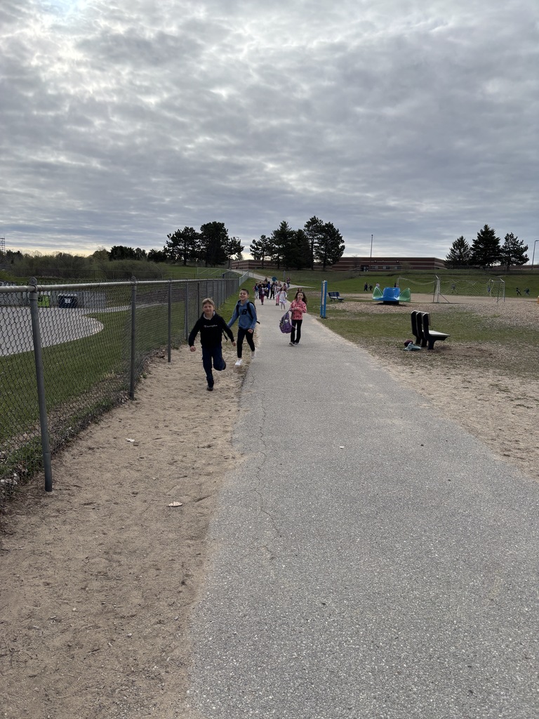 Children running and walking down a paved path outdoors next to a chain-link fence and a school playground.
