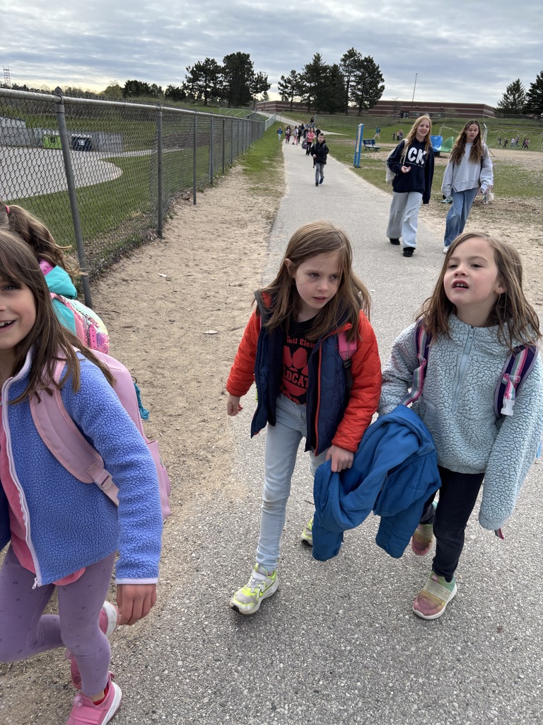 Elementary students with backpacks walking along an outdoor paved path next to a school playground and fence.