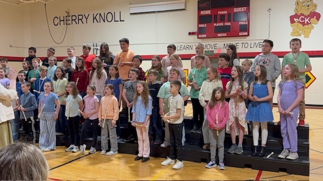 A group of children on a stage, possibly singing, in a gymnasium with a red wall.