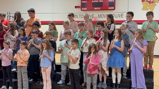 Group of children standing on stage, holding and playing flutes, in a room with red wall.