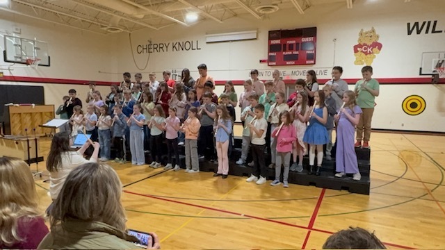 A group of children stand on stage in a gymnasium, dressed in various colored outfits, clapping and performing.