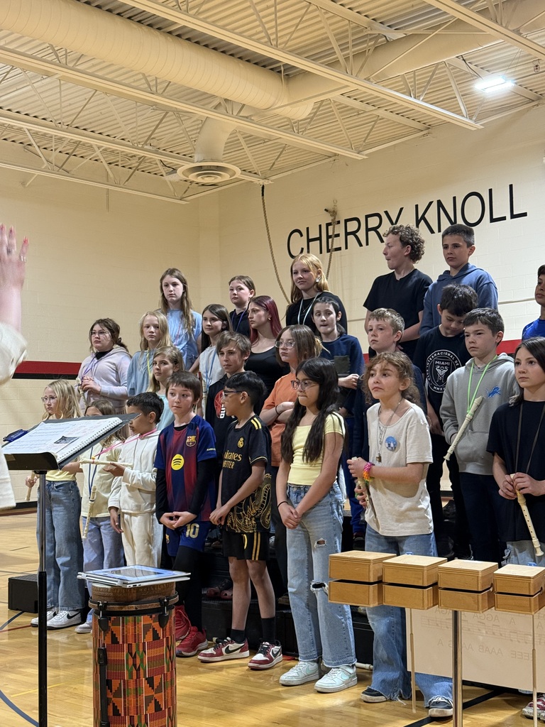 A group of children stand in a gymnasium, with some holding musical instruments, in front of a wall with "CHERRY KNOLL" written on it.