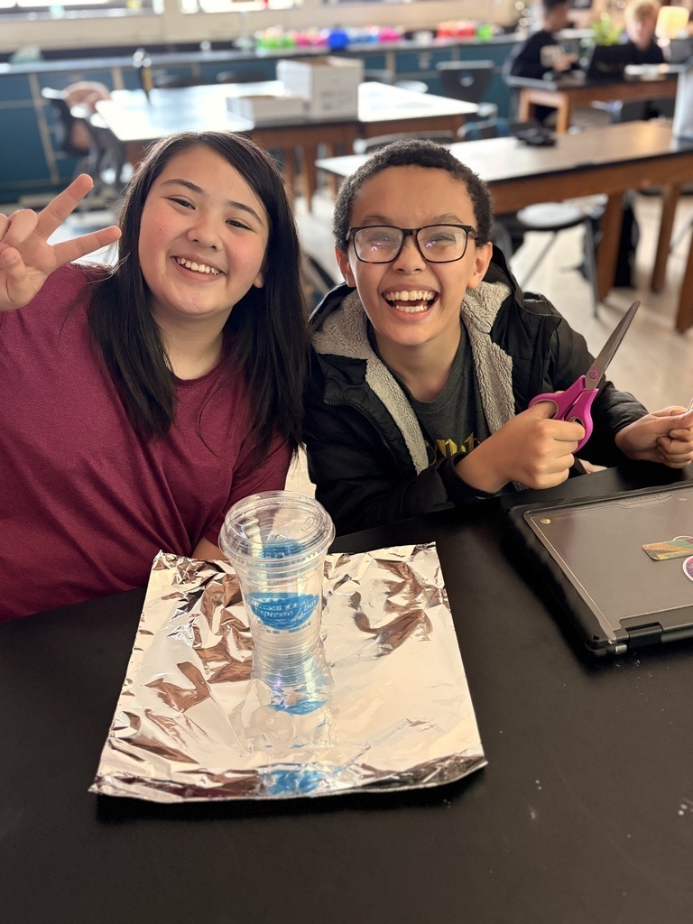 Two students sit at a table in a classroom, one holds scissors and the other poses with a peace sign.