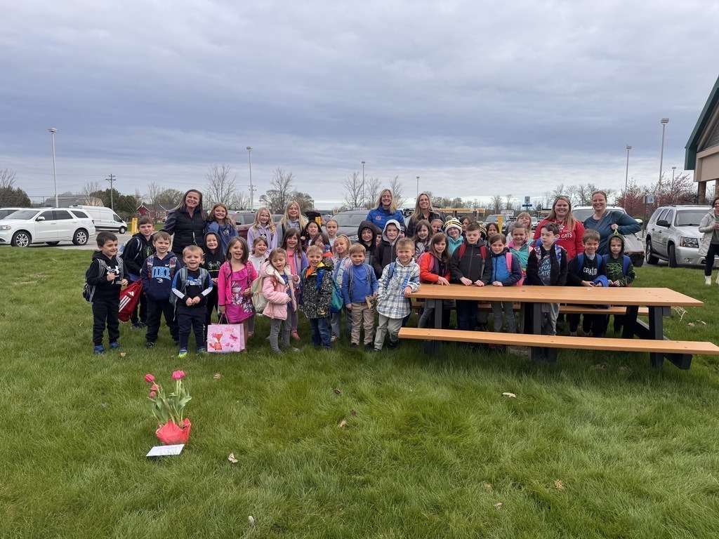 A group of children and adults gather near a picnic table on a grassy field with cars parked in the background.