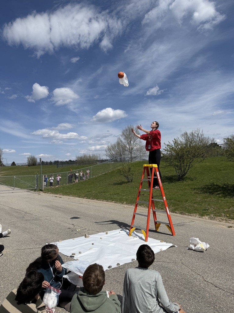 A student's egg drop project falls toward a target while a group of students observes the experiment.
