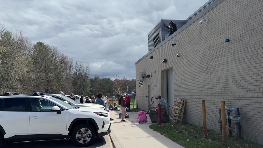 Exterior of a building with several cars parked outside. People are walking on a sidewalk. Trees in the background.