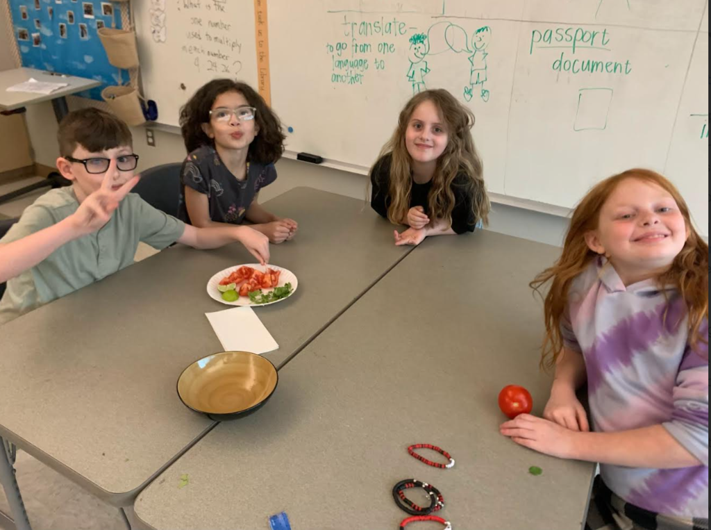 Four children sit around a table in a classroom. One child poses with a peace sign.