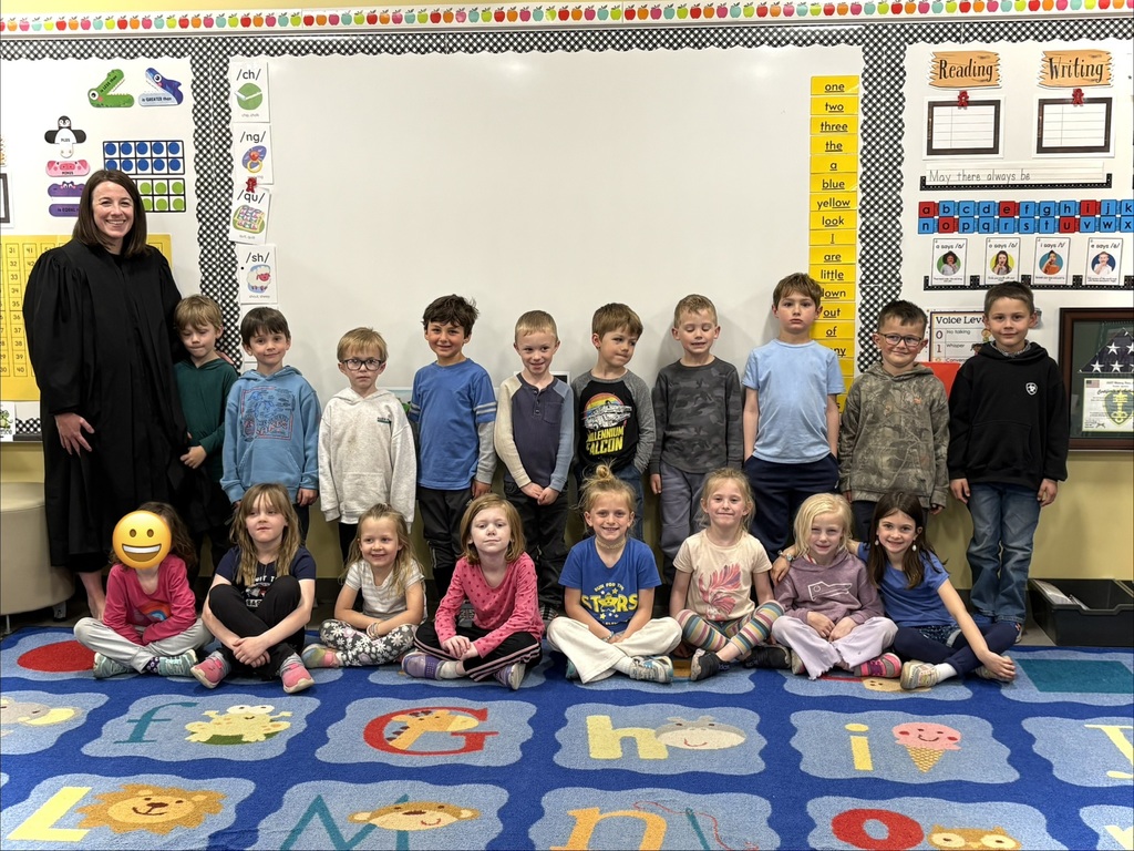 A group of children sitting on a floor in front of the classroom white board with Judge Whitten who is standing in the left corner back row. The floor has an alphabet carpet.